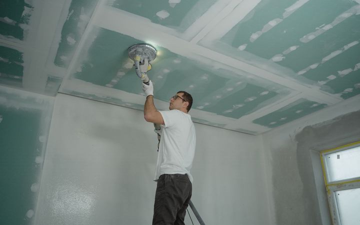 Worker on a stepladder sanding the taped seams of a drywall ceiling